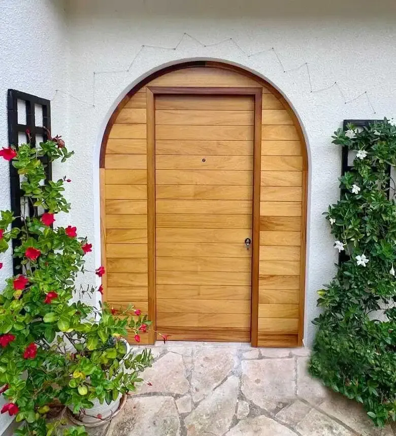 Arched iroko front door with horizontal boards in a white rendered wall framed by climbing plants