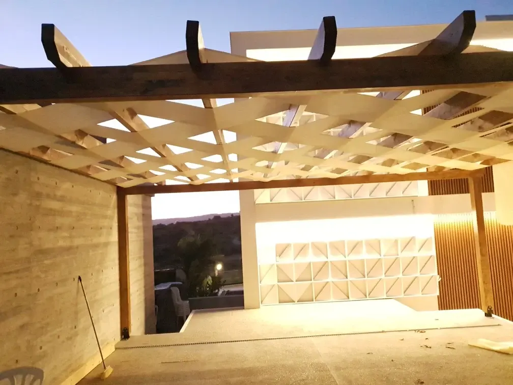Timber pergola with a woven shade canopy lit up at night against a board-formed concrete wall